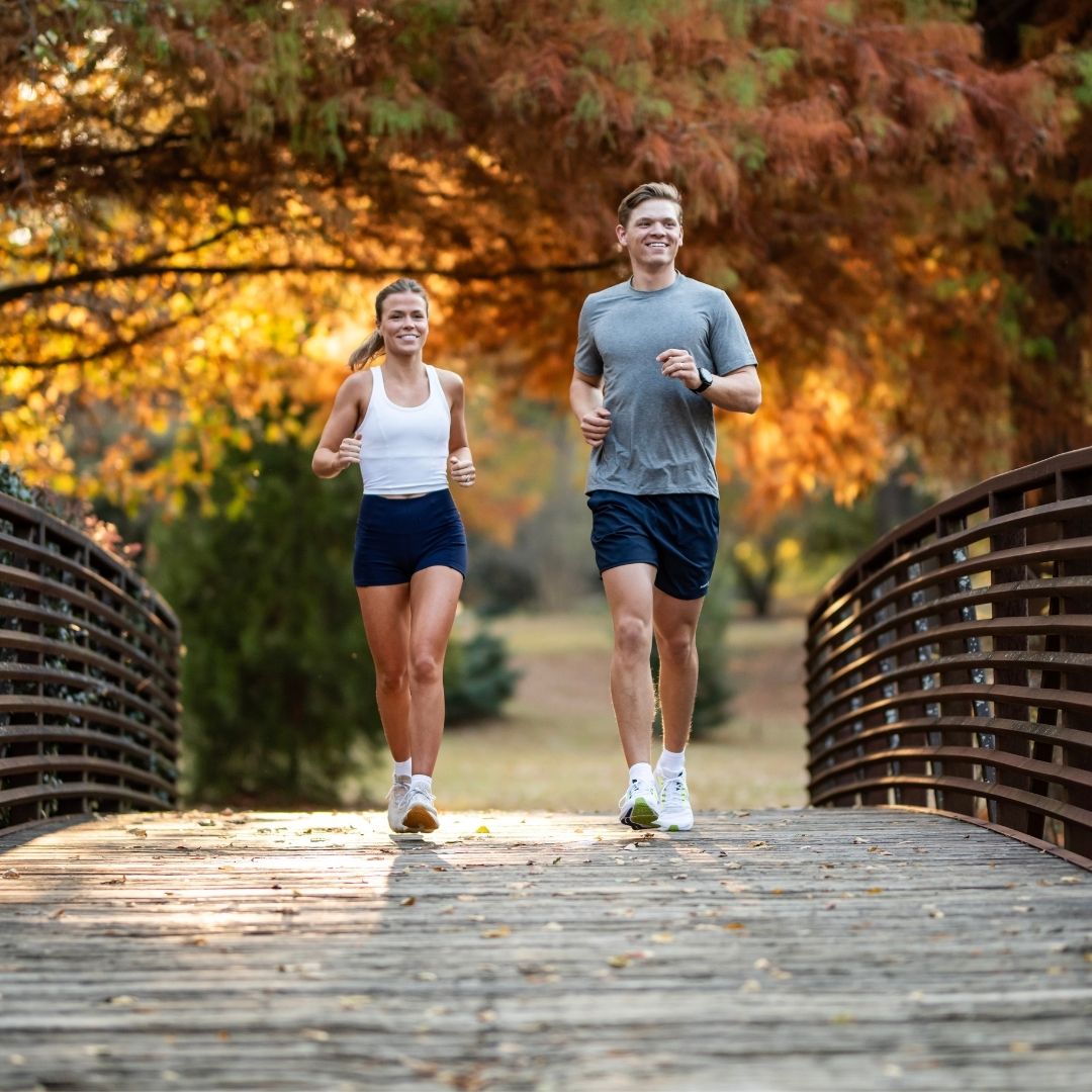 Two people running on a wooden path with autumn foliage in the background
