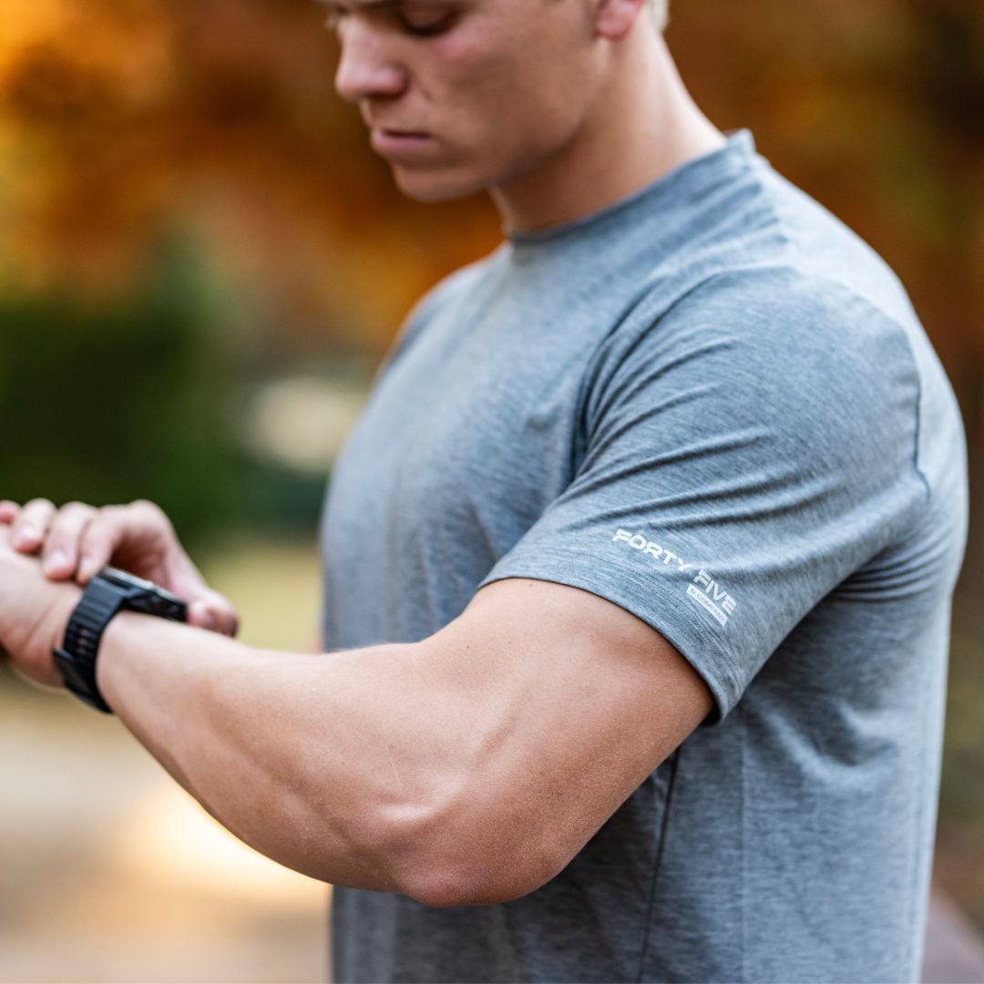Person wearing a gray t-shirt with a brand logo, looking at their watch outdoors.