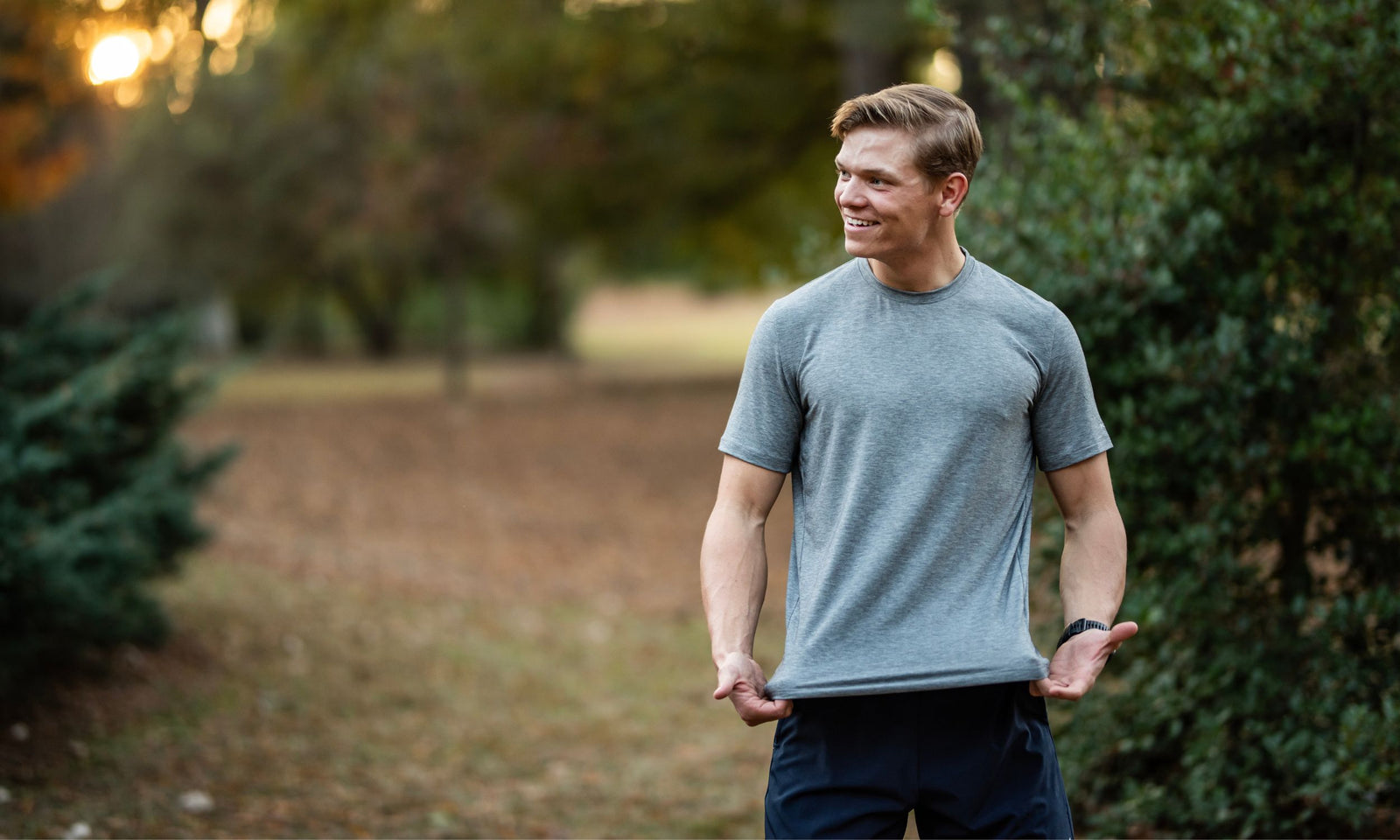 Man in a gray t-shirt standing outdoors with greenery in the background