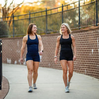 Two women walking outdoors on a sidewalk with a brick wall and metal railing in the background.