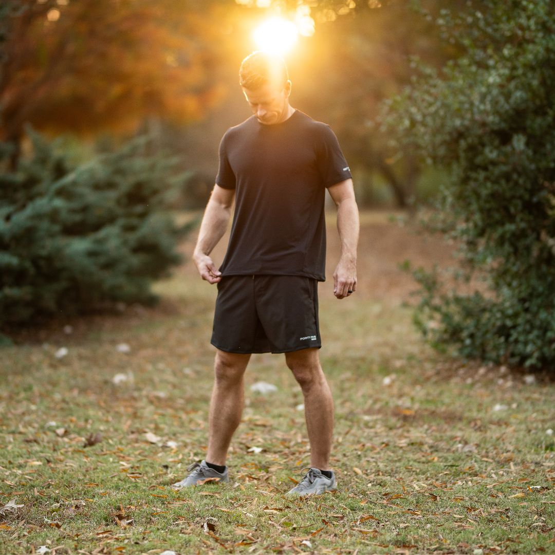 Man in black athletic wear standing in a park with trees and sunlight filtering through.